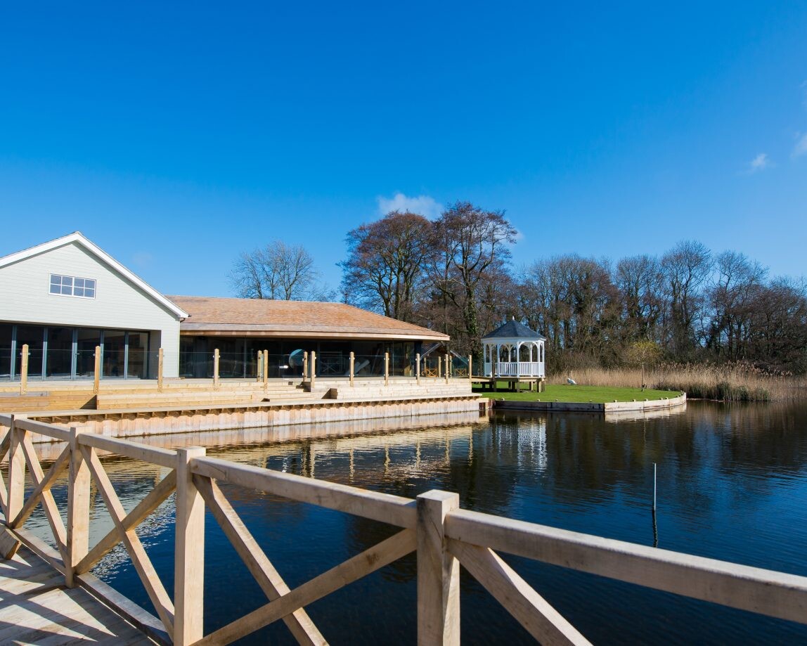 The Boathouse, a lakeside building with large windows, stands next to a wooden railing and walkway. A white gazebo graces a small platform by the water. The serene scene is framed by trees under a clear blue sky.