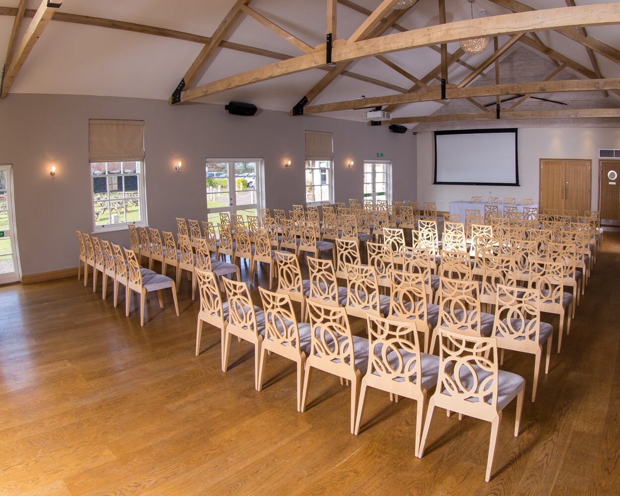 The Boathouse event hall boasts wood flooring and exposed beams, with rows of ornate wooden chairs facing a projector screen. Large windows flood the space with natural light, while three doors enhance its inviting atmosphere for memorable gatherings.
