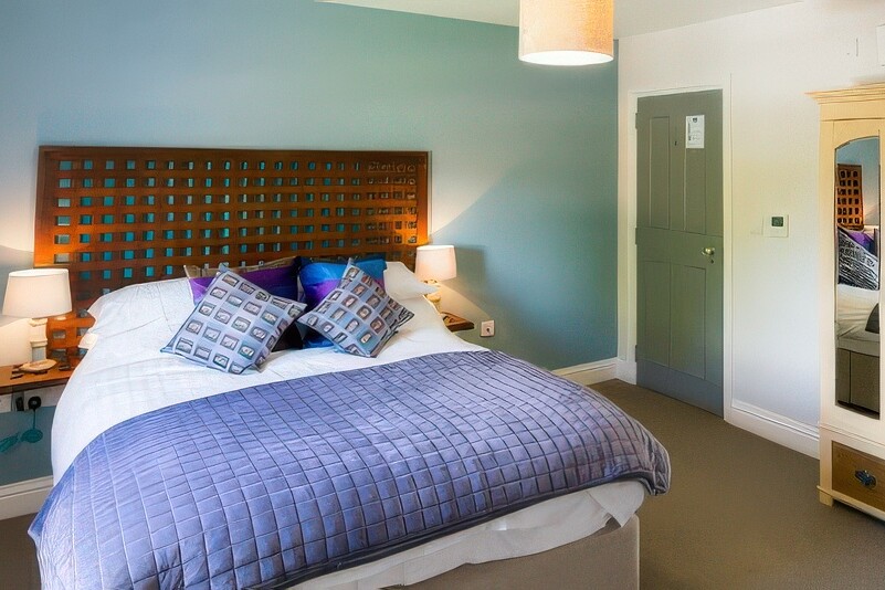 The cozy bedroom at The Boathouse features a double bed adorned with a blue quilt and patterned pillows. Two lamps rest on bedside tables beside a wooden wardrobe with mirrored doors. The blue and white walls complement the lit ceiling lamp hanging above.
