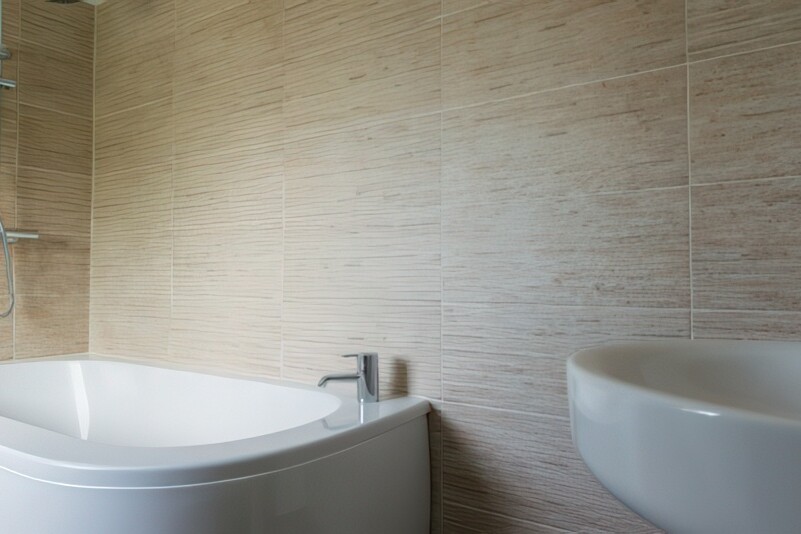 A modern bathroom at The Boathouse showcases beige tiled walls, a sleek white bathtub with a glass shower enclosure, and a mounted silver showerhead. The minimalist decor is completed by a white sink with a silver faucet, embodying clean and elegant design.