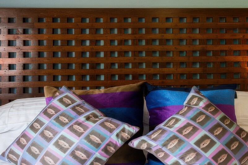 A cozy bedroom at The Boathouse features a wooden lattice headboard, adorned with two lit bedside lamps. The neatly made bed boasts white sheets and decorative pillows in vibrant blue and brown patterns, creating a warm and inviting atmosphere.