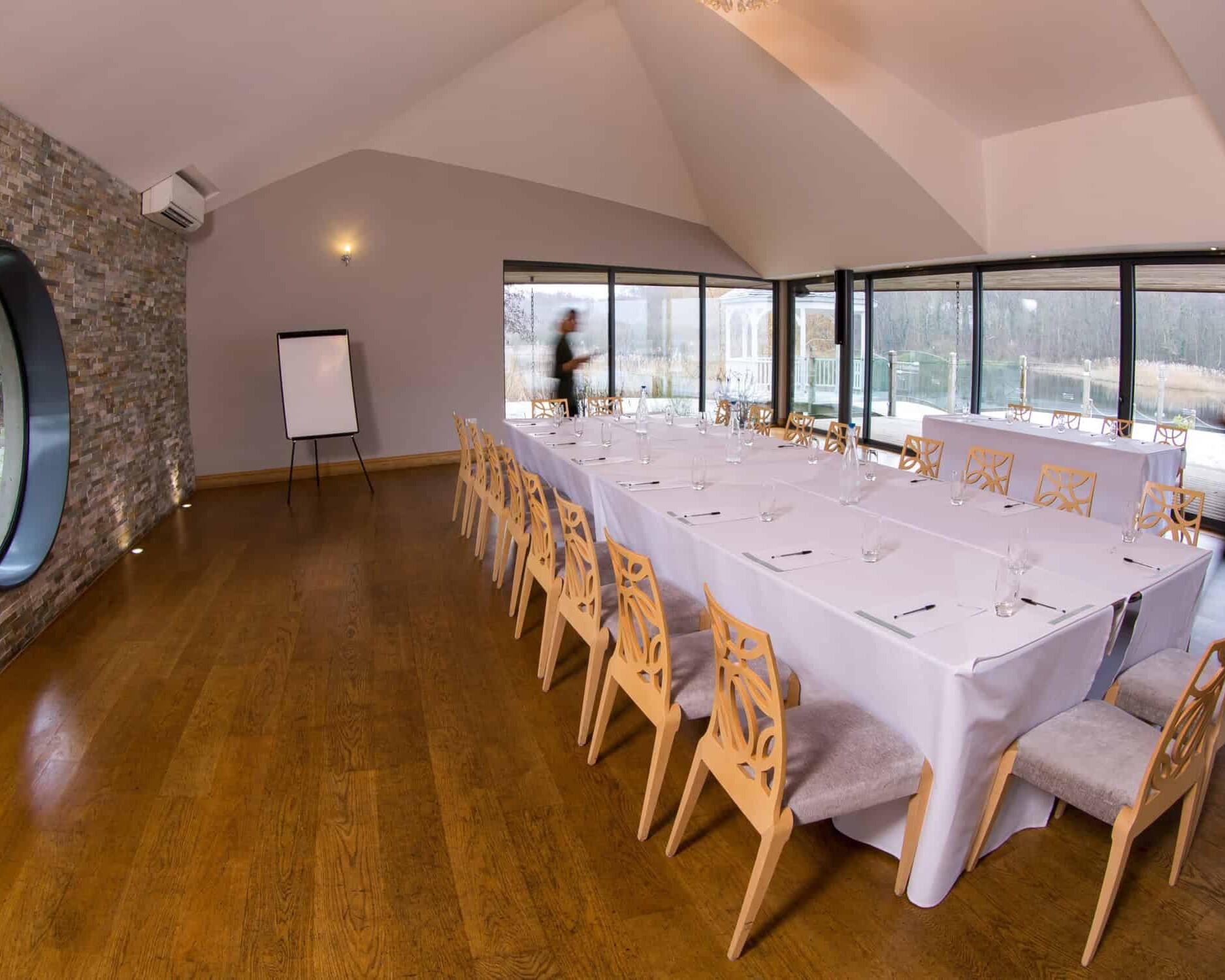 A spacious conference room with a rectangular setup of tables draped in white cloths, surrounded by wooden chairs. There's a flip chart near a circular window on a brick wall. Large glass windows on one side showcase an outdoor view.