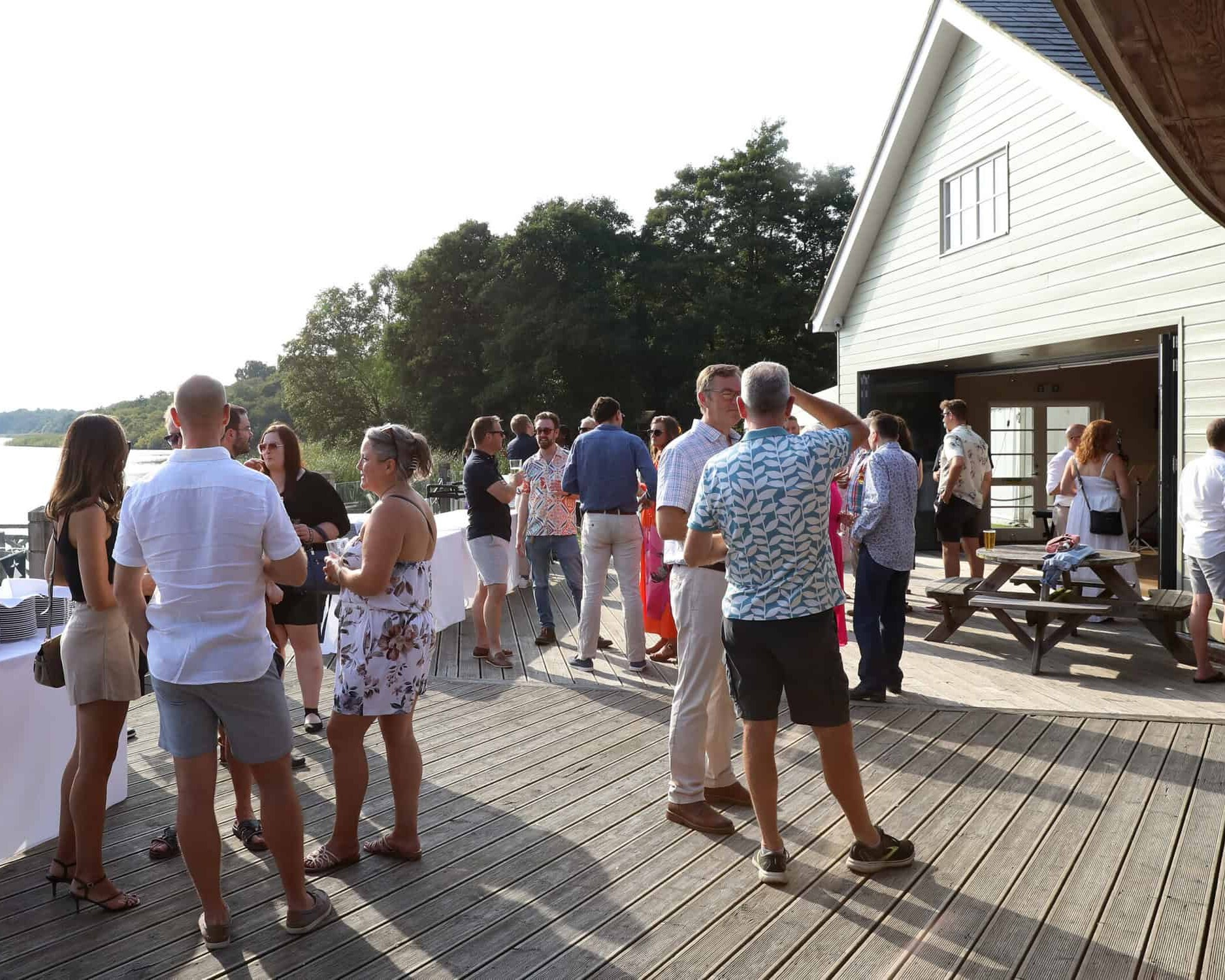 A group of people socializing on a wooden deck beside a large lake house. They are engaged in conversation and enjoying a sunny day. A table with plates is set up on the left, and trees and water are visible in the background.