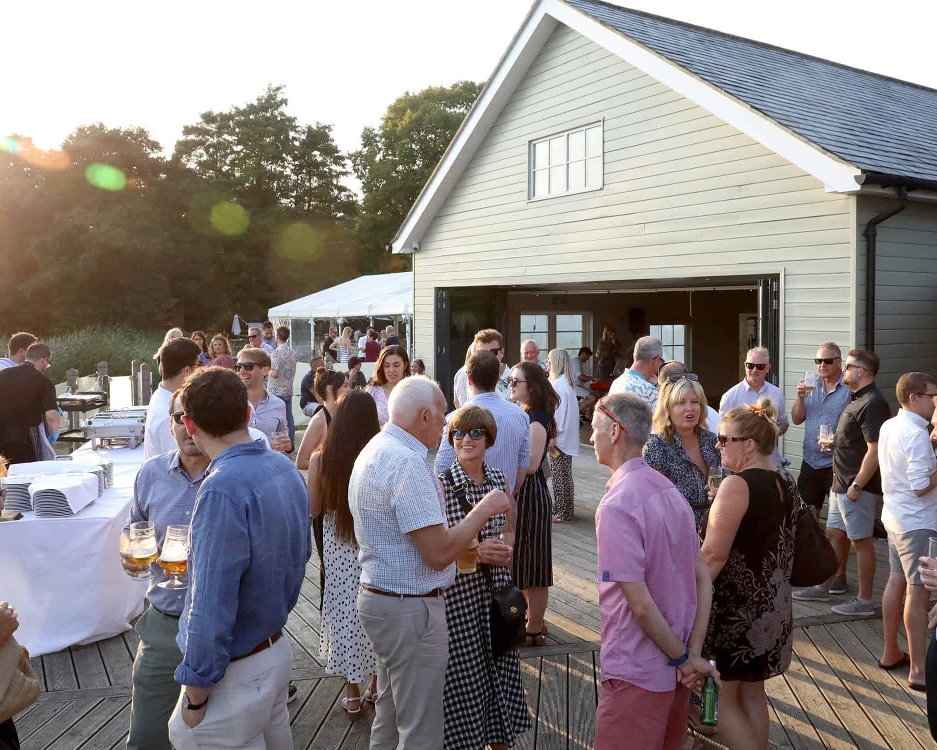 A diverse group of people socializing at an outdoor event on a wooden deck, near a light green building. They are dressed in summer attire, holding drinks, and engaged in conversation. The sun is shining, creating a warm and lively atmosphere.