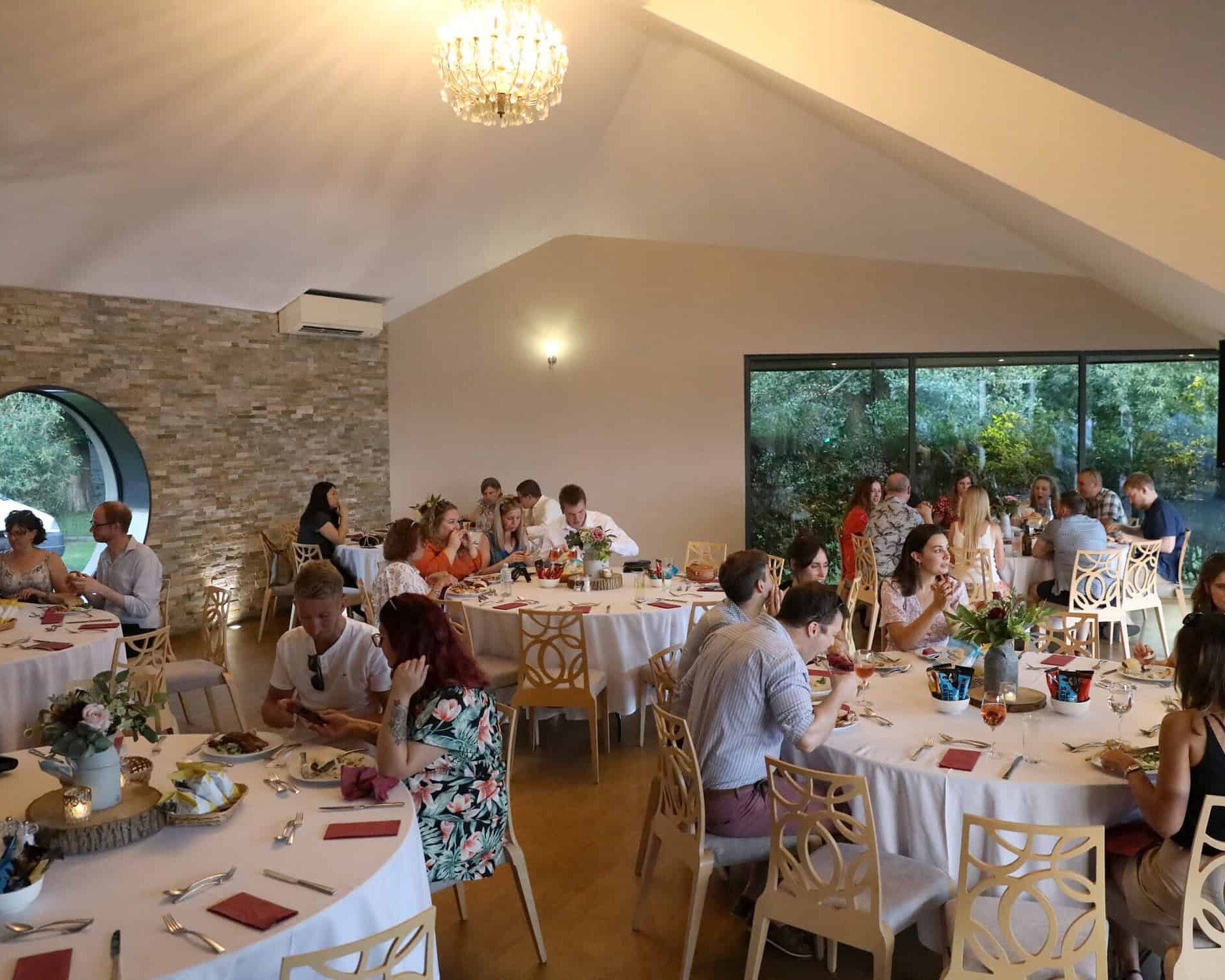 People are gathered at round tables in an elegant room with a high ceiling and large windows. The tables are set with white tablecloths, floral centerpieces, and dining settings. Some guests are seated while others stand and converse.