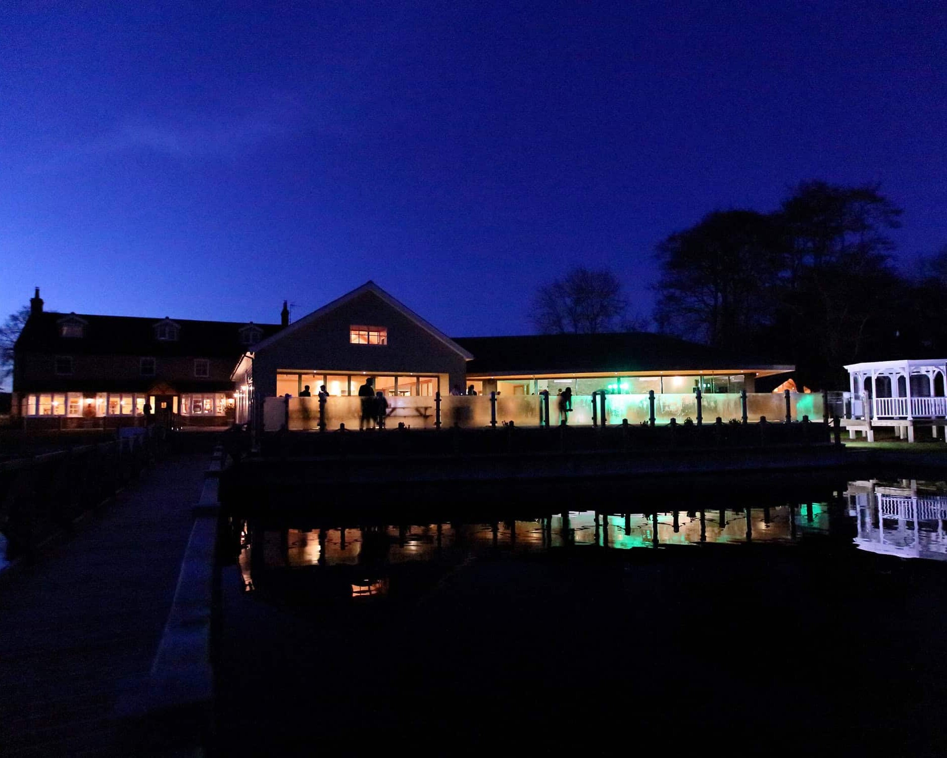 A nighttime scene of The Boathouse by a lake, its lights shimmering in the water. The indigo sky casts trees in silhouette, while a white gazebo stands elegantly to the right&mdash;perfect for private hire events or intimate functions.