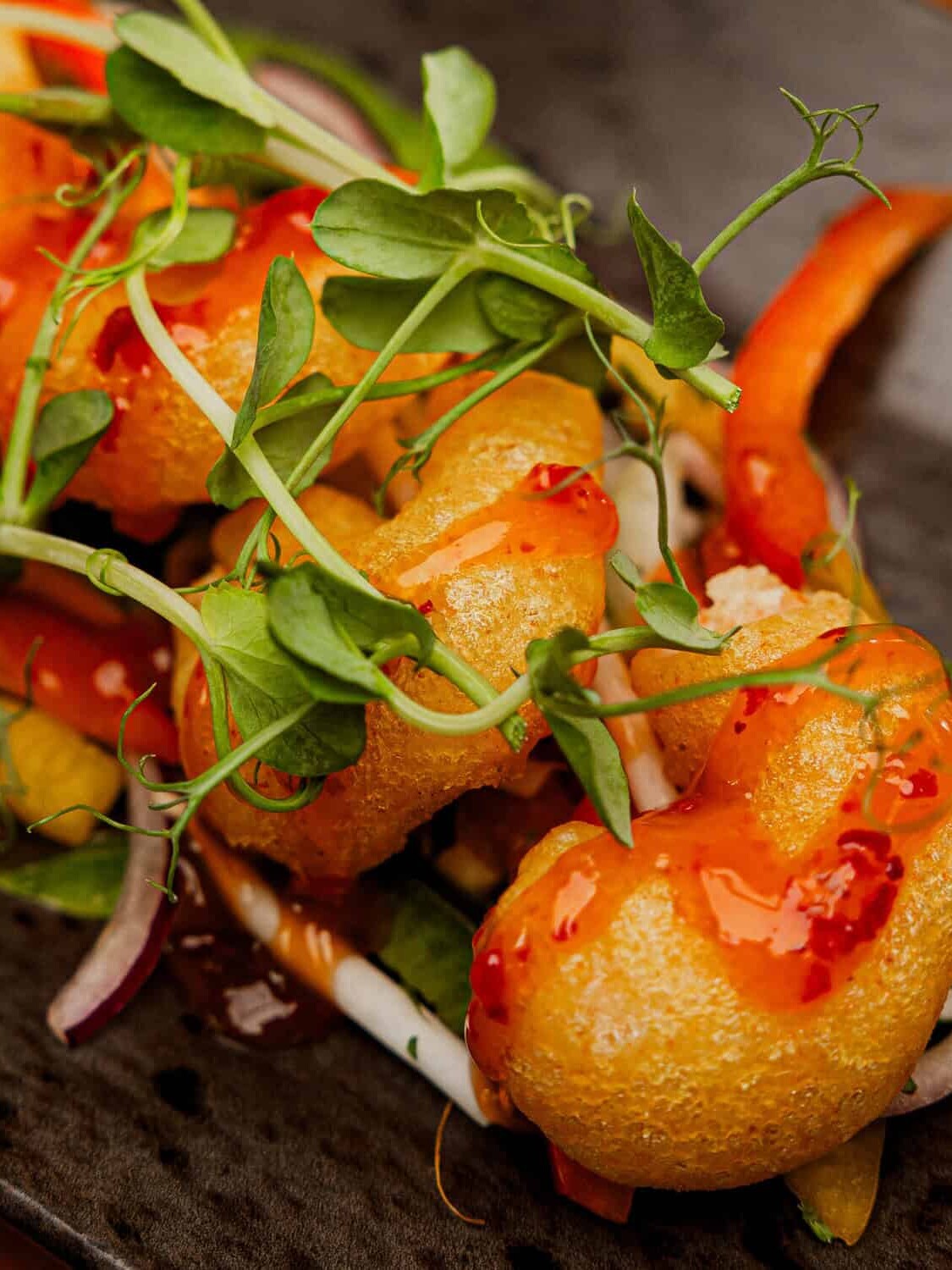 A close-up of crispy tempura shrimp drizzled with sweet chili sauce, garnished with microgreens, served on sautéed vegetables with a lime wedge on a dark plate.