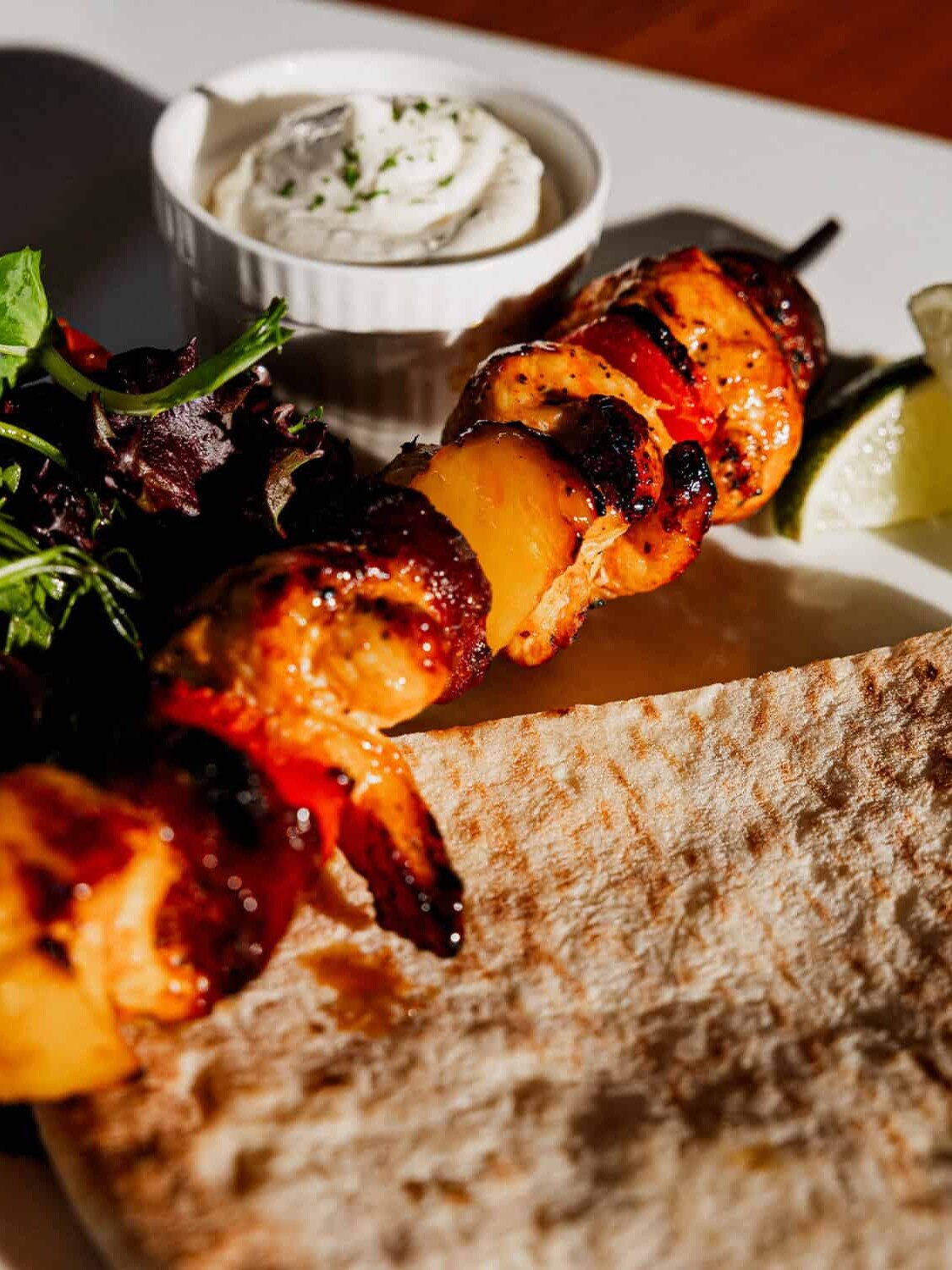 A plate with grilled chicken and vegetable skewers, flatbread, mixed greens, a lime wedge, and a small dish of creamy dip. Sunlight highlights the food on a white rectangular plate.