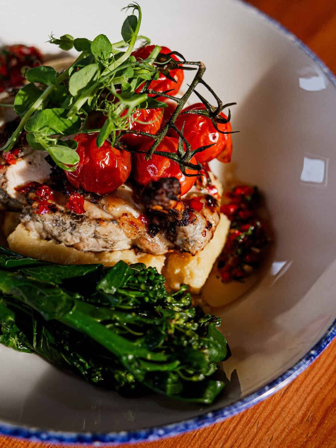 A white bowl with grilled meat topped with roasted cherry tomatoes and fresh greens, served with broccolini and a red sauce, on a wooden table.