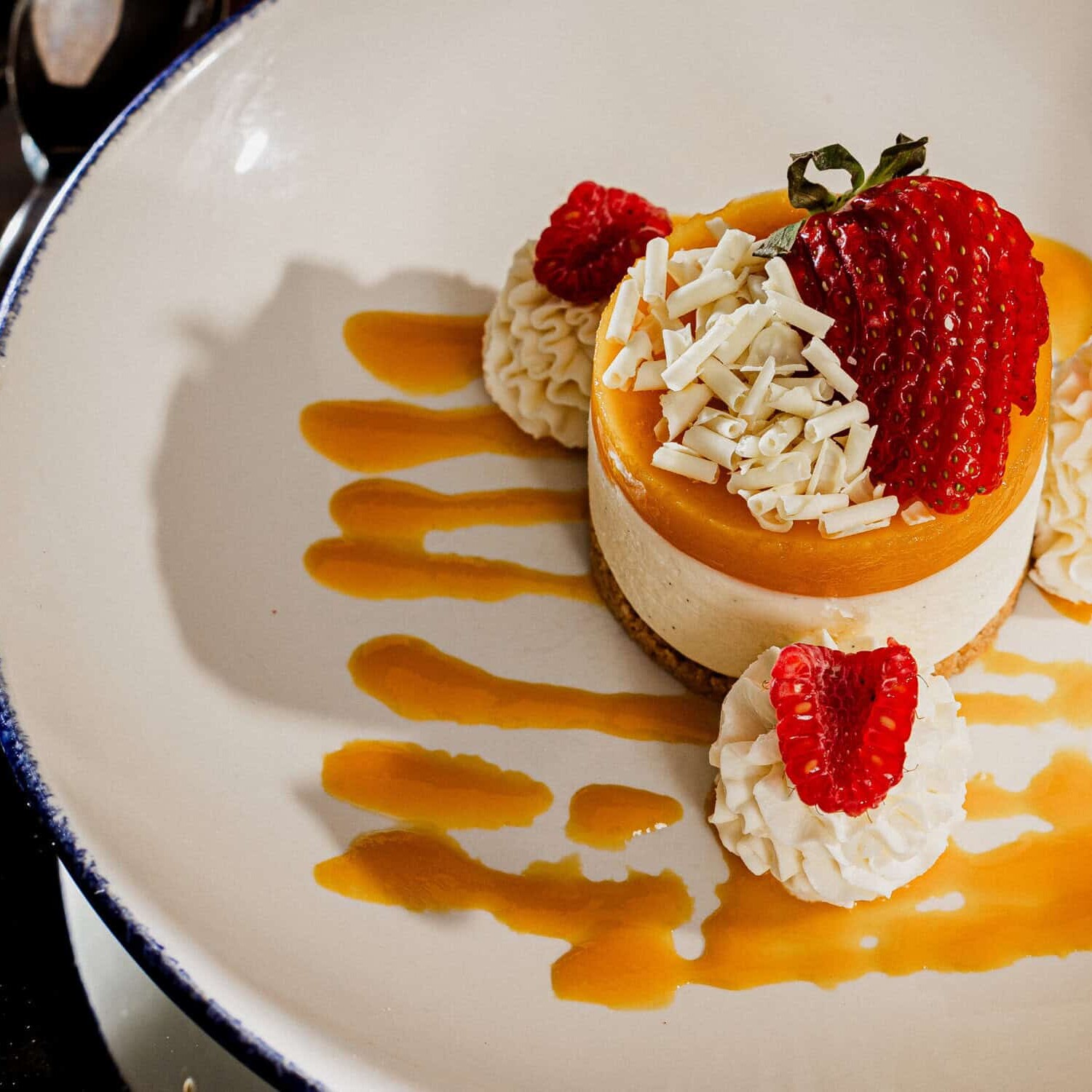 A round cheesecake topped with a strawberry, white chocolate shavings, and raspberries, sits on a white plate with caramel sauce and whipped cream, beside a fork and knife.