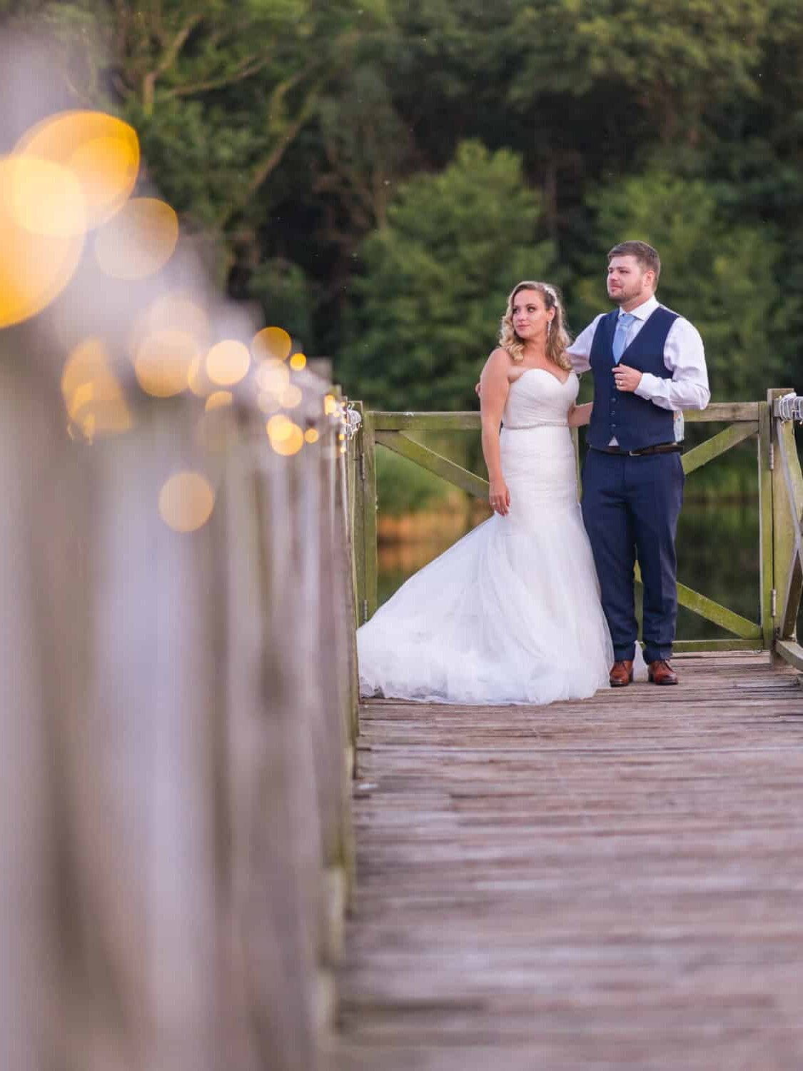 A bride in a white gown and a groom in a vest stand on a wooden bridge decorated with string lights. The background features a lush green forest and a calm lake, creating a serene and romantic atmosphere.