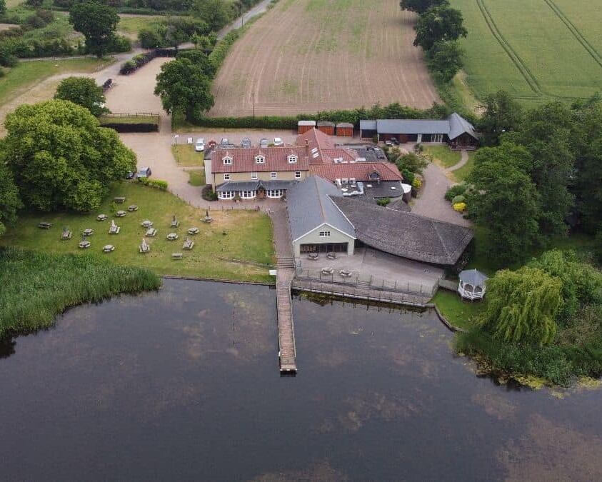 Aerial view of a lakeside property with a main building, outdoor seating area, and a small dock extending into the water. The property is surrounded by trees and fields, with pathways leading to various parts of the estate.