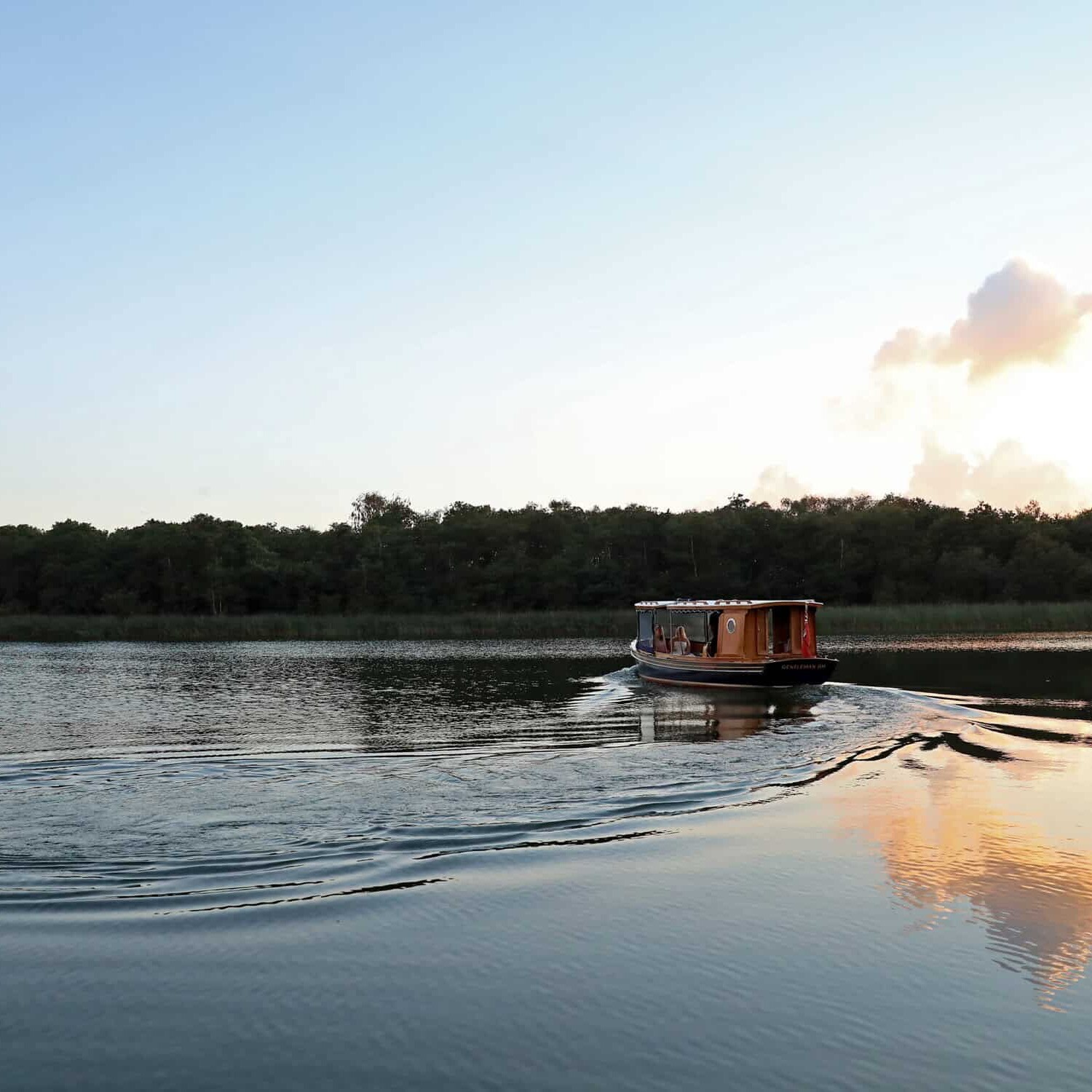 A small boat travels on a calm lake with ripples trailing behind. The sky is partially cloudy with a subtle sunrise or sunset glow, and lush greenery lines the horizon.